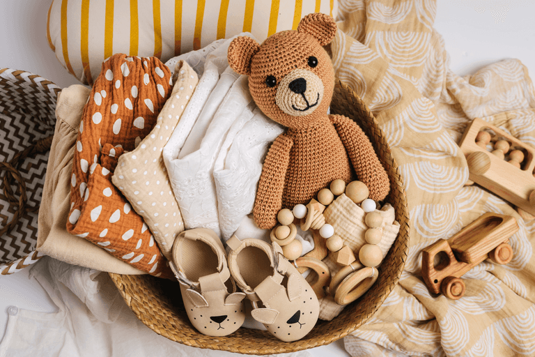 Basket of baby registry gifts: clothes, a knitted teddy bear, teething toys and soft shoes
