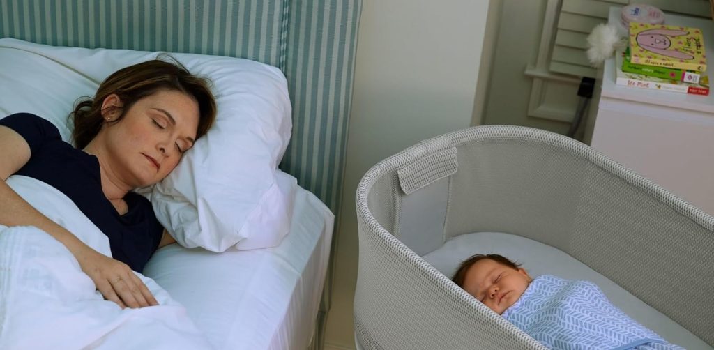 Parent asleep in bed next to a bassinet where the baby sleeps on their back — room-sharing without bed-sharing