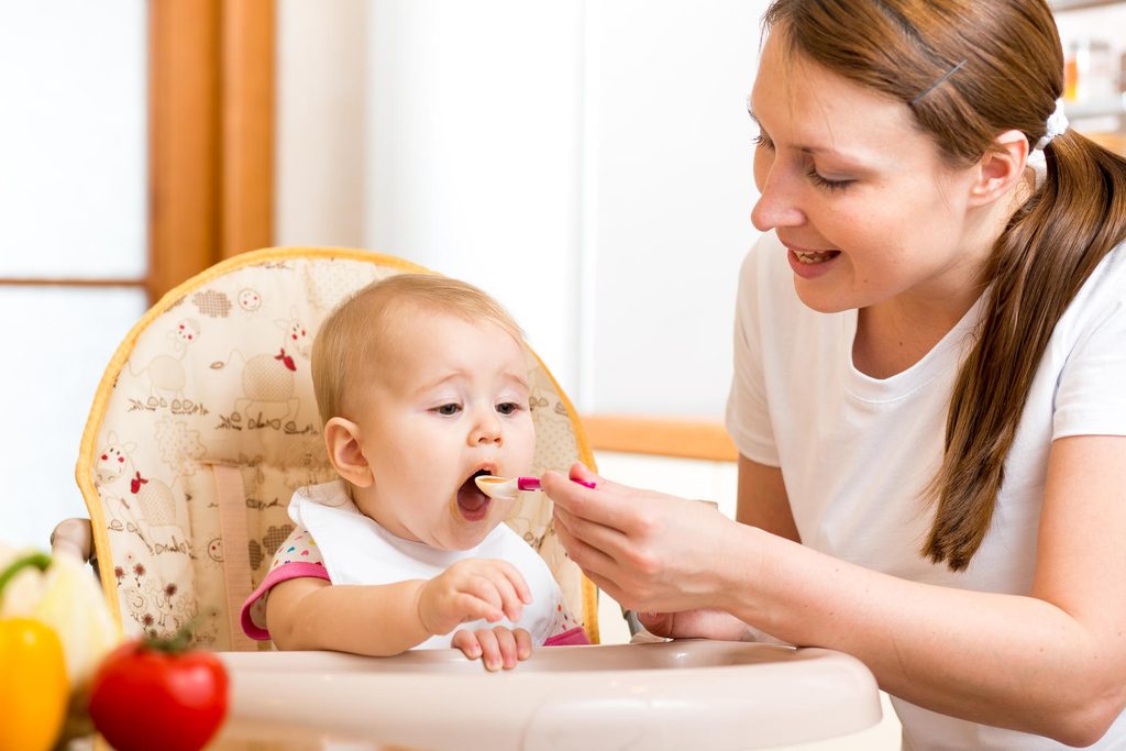 Mother spoon-feeding her baby in a high chair with fresh vegetables nearby